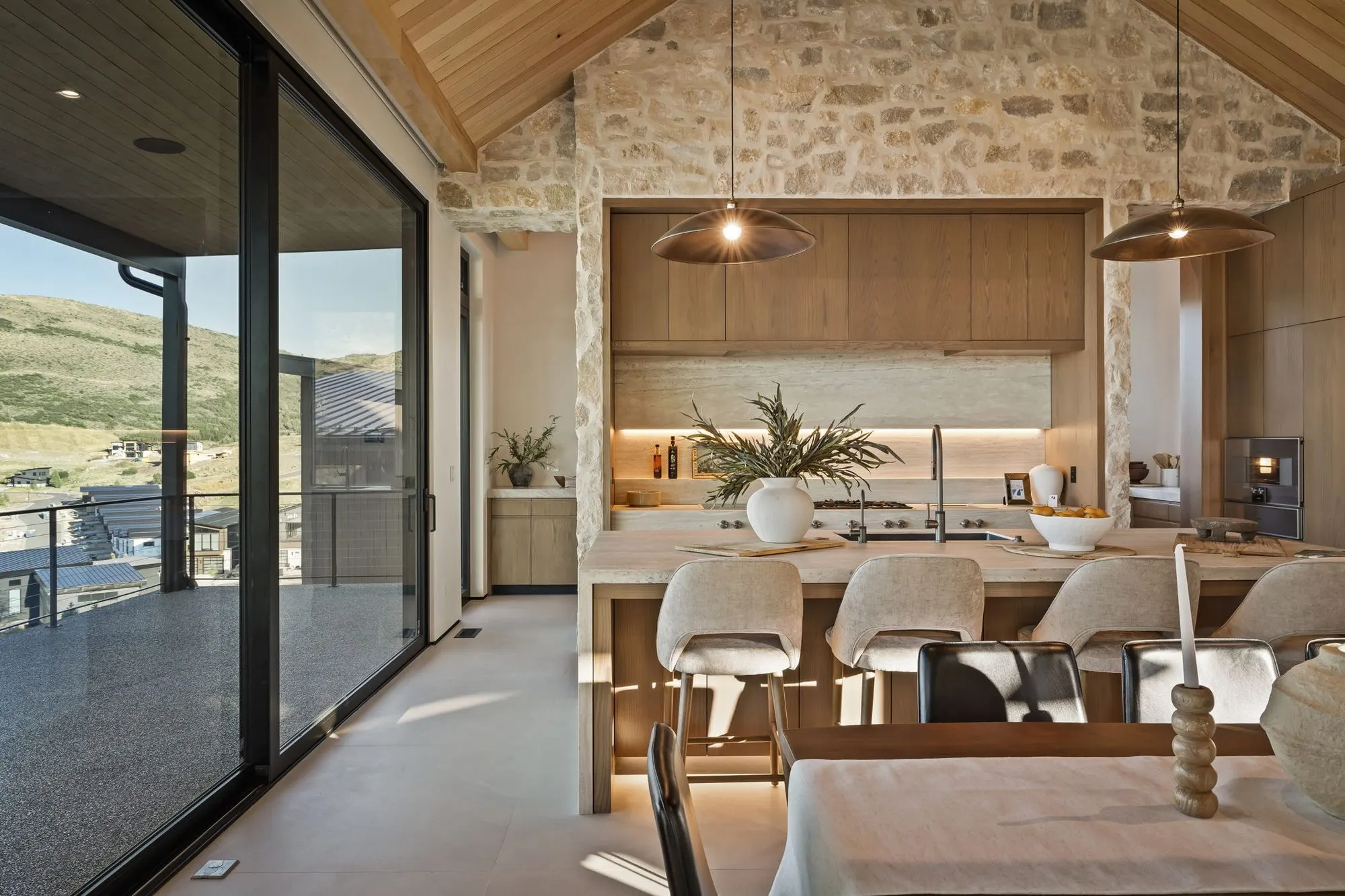Lumen — kitchen island and counter seating against the stone wall, glass sliders to the deck