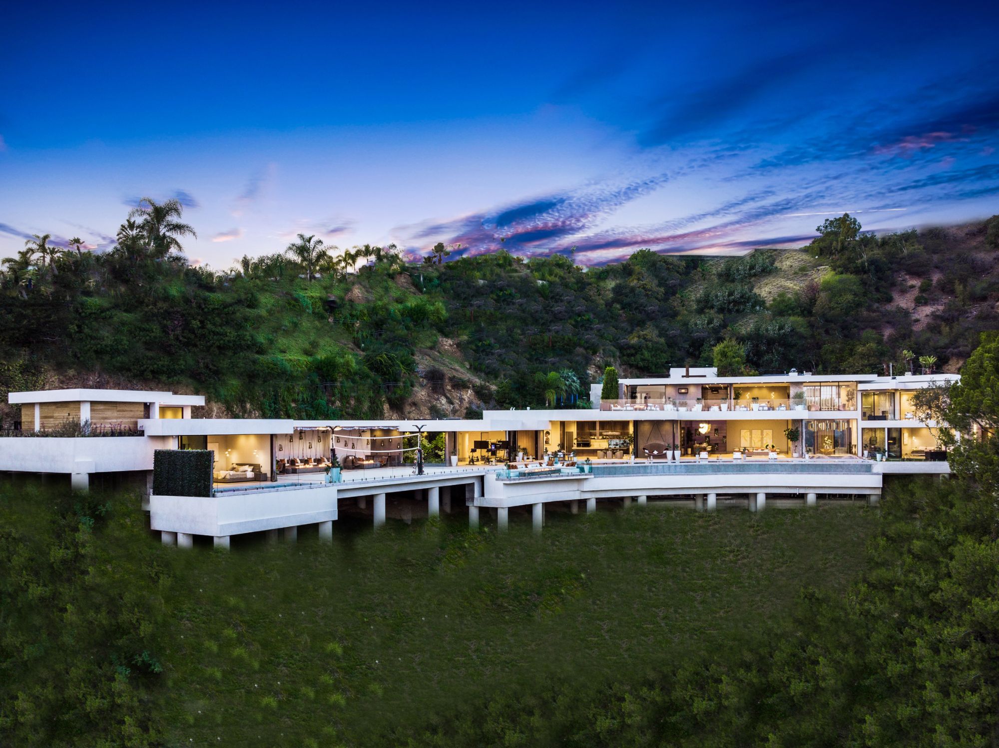 The Flicker Estate at dusk from across the canyon — the curved house stepping along the hillside, the pool deck cantilevered out, the whole structure standing on concrete piles