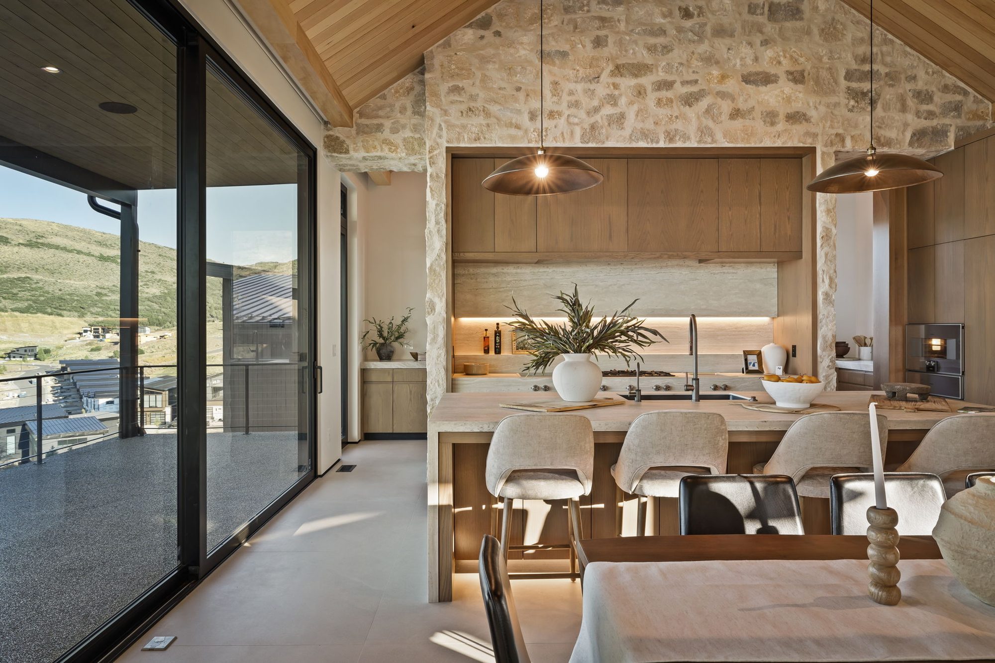 Lumen — kitchen island and counter seating against the stone wall, glass sliders to the deck